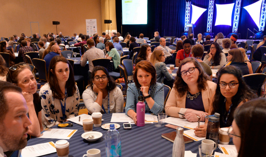UMBC students, faculty, and staff sit around a table and engage in a group conversation at a crowded conference.